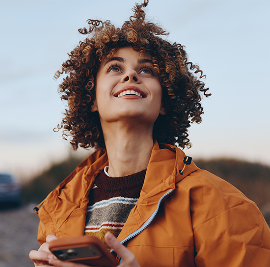 Woman with curly hair smiling outdoors while holding a smartphone, representing patients in the San Diego community booking appointments